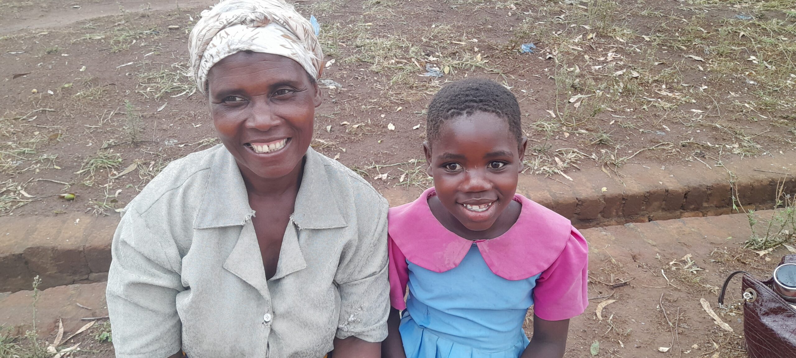 The image shows a smiling woman and a young girl sitting side by side outdoors. The woman is wearing a headscarf and a light-colored shirt, while the girl is dressed in a blue dress with a pink collar. The background is a dirt ground with sparse vegetation.